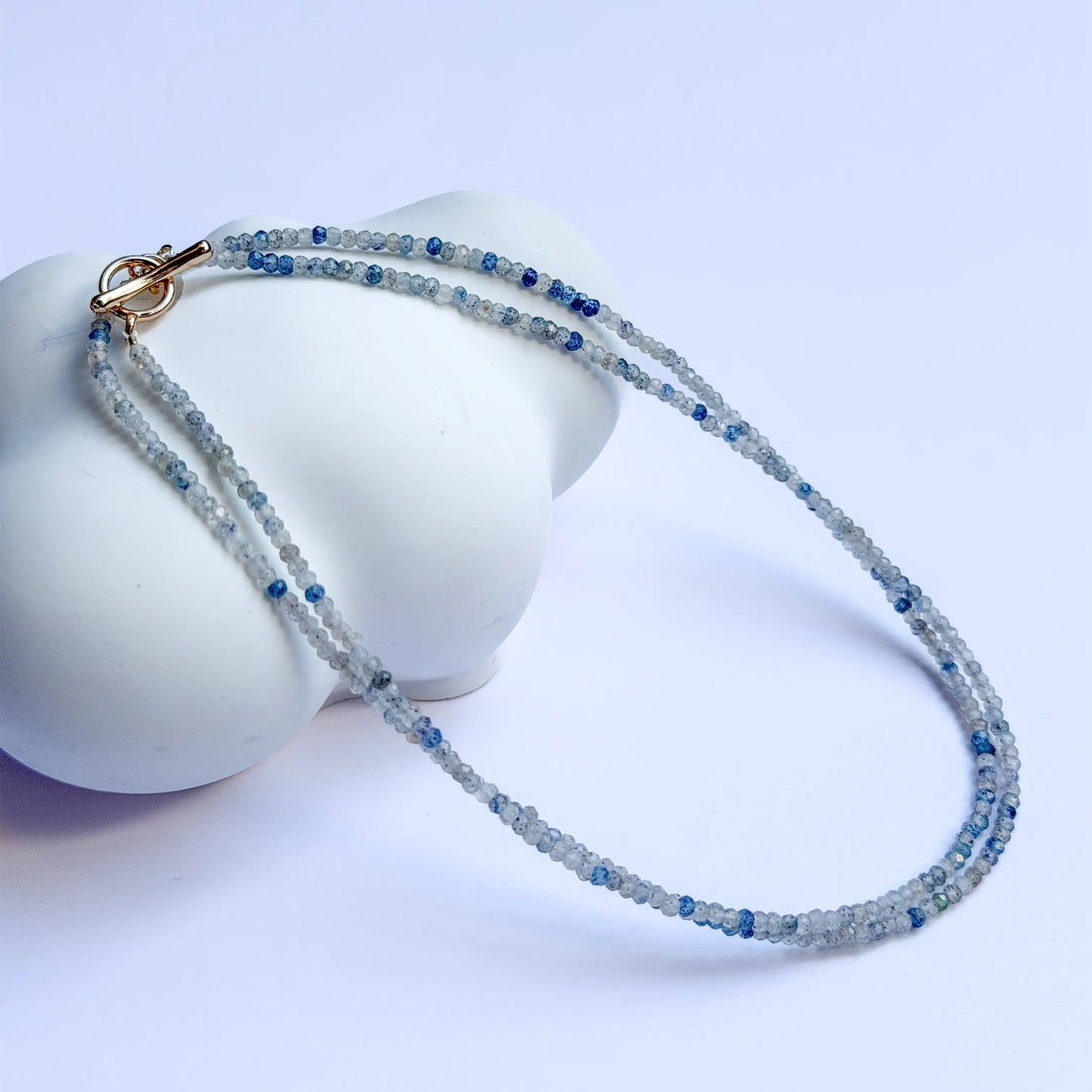 K2 Mineral Necklace with blue and clear beads on a white stand against a light purple background
